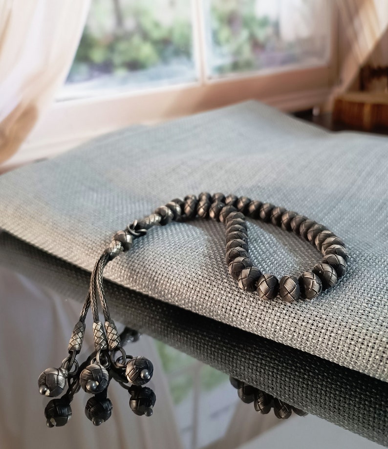 May include: A silver-toned beaded necklace with a woven design. The necklace is resting on a grey textured surface. The necklace has a tassel of small, decorative silver balls.