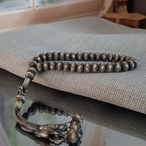 May include: A silver-coloured prayer bead bracelet featuring woven beads and a decorative tassel. The round beads are evenly spaced, with a textured surface. The tassel and connecting elements are also silver-coloured, enhancing the bracelet's overall appearance.