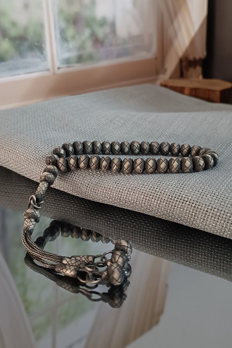 May include: A silver-coloured prayer bead bracelet featuring woven beads and a decorative tassel. The round beads are evenly spaced, with a textured surface. The tassel and connecting elements are also silver-coloured, enhancing the bracelet's overall appearance.