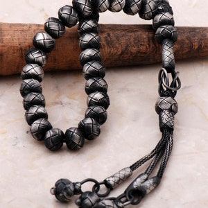 May include: A silver-toned beaded necklace with a woven pattern on each bead. The necklace has a tassel at the end and is displayed on a wooden dowel. The beads are approximately 1 cm in diameter.