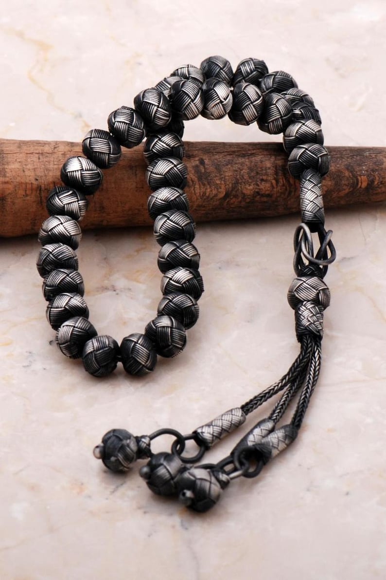May include: A silver-toned beaded necklace with a woven pattern on each bead. The necklace has a tassel at the end and is displayed on a wooden dowel. The beads are approximately 1 cm in diameter.