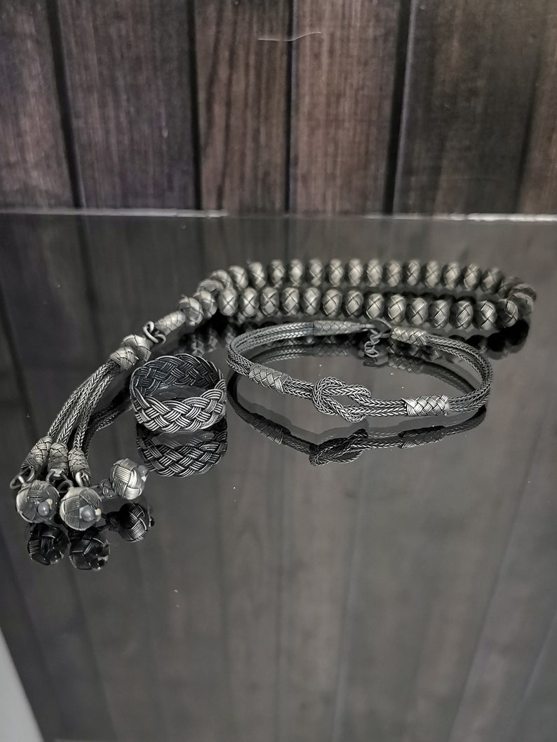 May include: A set of silver-coloured jewellery including a braided ring, a bracelet with a knot design, and a beaded strand with tassel ends. The items are displayed on a reflective surface.