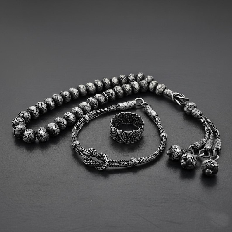 May include: A collection of silver-coloured jewellery, featuring a beaded strand, a bracelet with a knot design, and a woven ring. The items are displayed on a dark surface, highlighting the intricate details.
