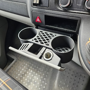 May include: Black car console cup holder with two circular openings and a rectangular storage area. The console also features a cigarette lighter, a hazard light button, and two USB ports. The interior has a patterned floor mat.