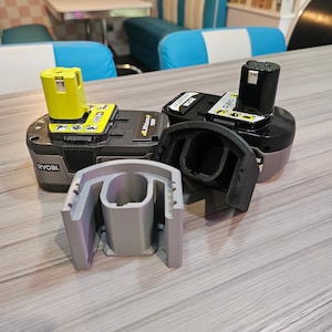 Op de afbeelding: Twee grijze en zwarte accu's voor een Ryobi-gereedschap. De accu's liggen op een houten tafel met een grijze 3D-geprinte houder.