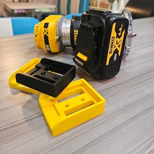 May include: A yellow and black DeWalt XR 18V 4.0 Ah battery and a yellow and black DeWalt XR 18V router with a black base and a clear plastic guard. The router and battery are sitting on a light brown wooden surface. There are two yellow plastic bases and one black plastic base on the surface.