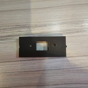 May include: A black plastic wall plate with a rectangular cutout and two small holes.