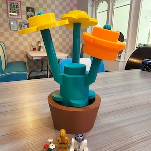 Puede incluir: Una maceta de flores impresa en 3D con tres flores amarillas y una flor naranja. La maceta es marr&oacute;n y las flores est&aacute;n en tallos de color turquesa. Dos peque&ntilde;as figuras de acci&oacute;n est&aacute;n delante de la maceta, una es dorada y la otra es blanca y azul.