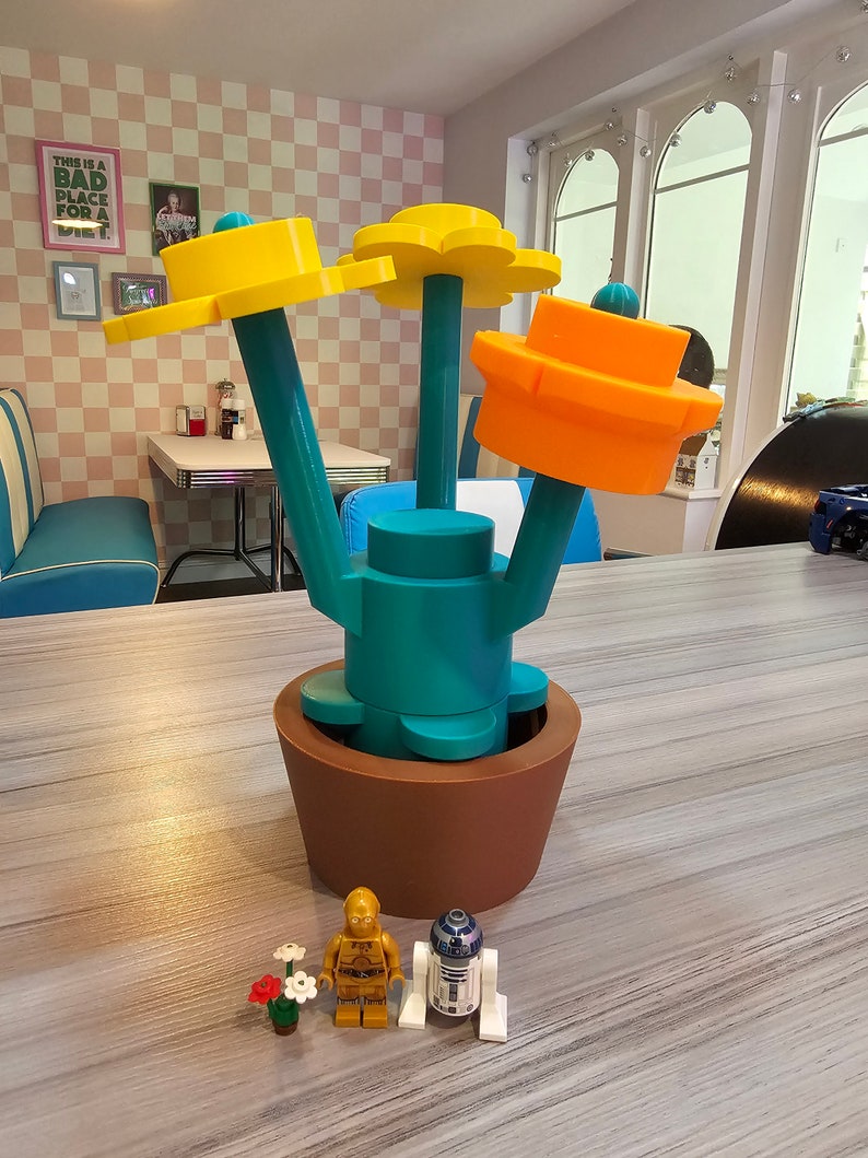 Puede incluir: Una maceta de flores impresa en 3D con tres flores amarillas y una flor naranja. La maceta es marr&oacute;n y las flores est&aacute;n en tallos de color turquesa. Dos peque&ntilde;as figuras de acci&oacute;n est&aacute;n delante de la maceta, una es dorada y la otra es blanca y azul.