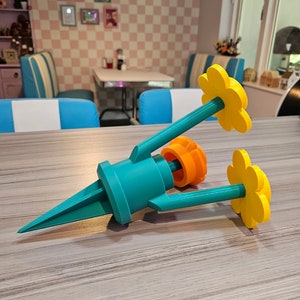 May include: A teal blue and orange plastic garden stake with three yellow flower-shaped decorations. The stake is designed to hold a small plant or flower.