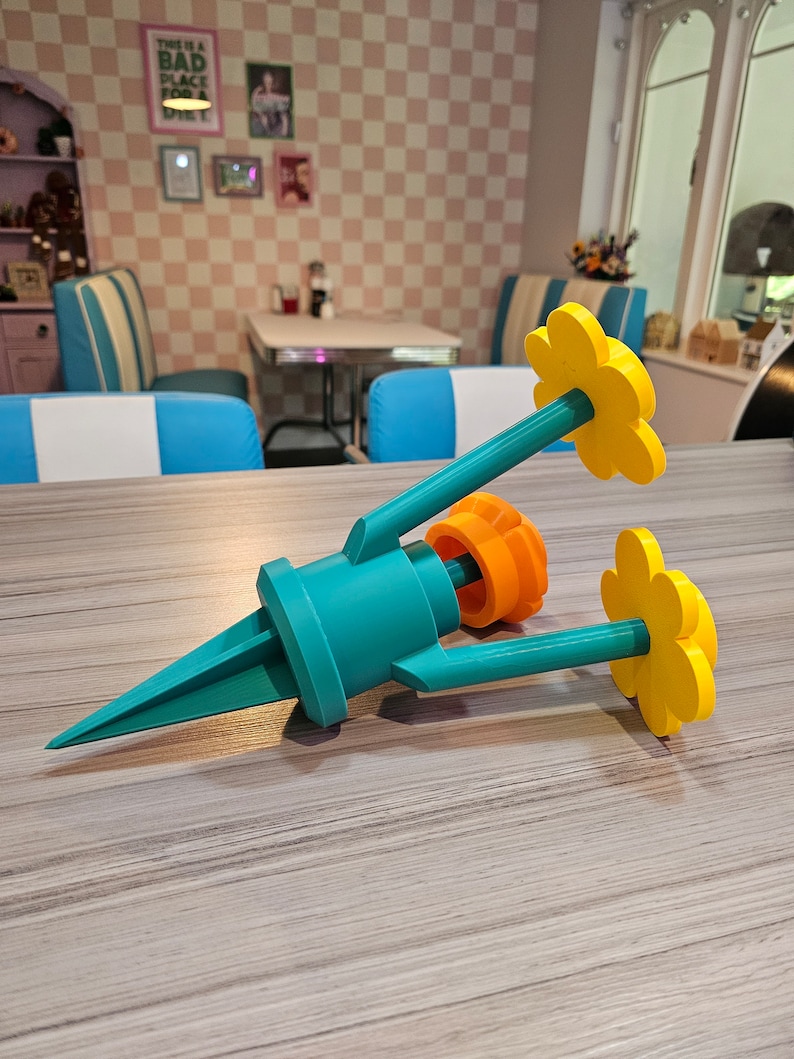 May include: A teal blue and orange plastic garden stake with three yellow flower-shaped decorations. The stake is designed to hold a small plant or flower.