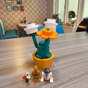 Puede incluir: Una maceta amarilla con una planta verde con forma de cactus con tres copas de flores blancas y una flor naranja. Dos peque&ntilde;as figuras, un robot dorado y un robot blanco, est&aacute;n de pie frente a la planta.