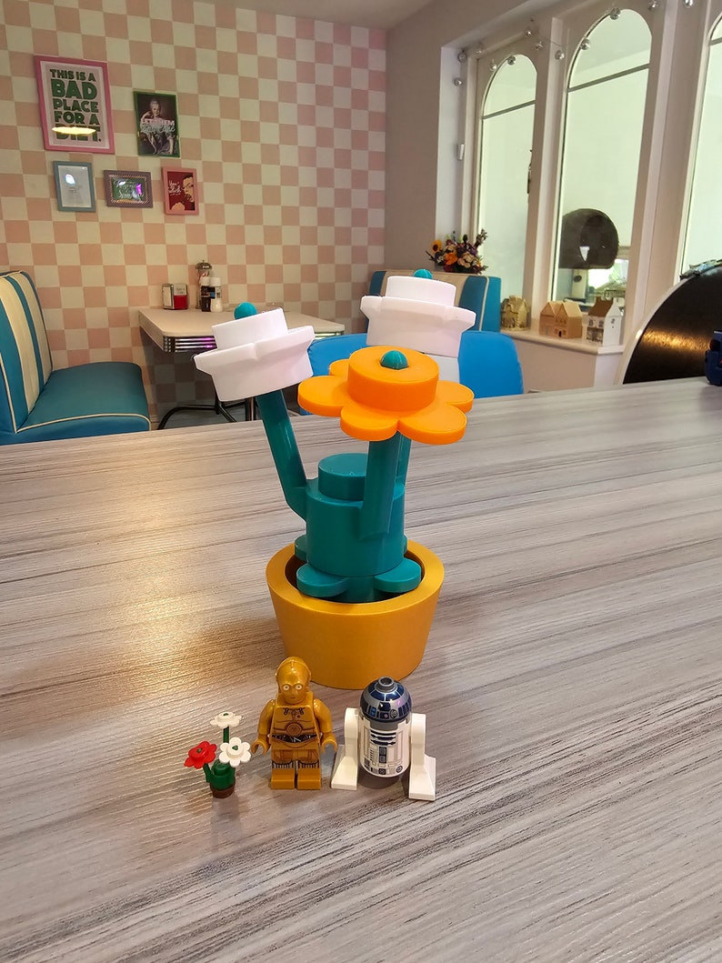 Puede incluir: Una maceta amarilla con una planta verde con forma de cactus con tres copas de flores blancas y una flor naranja. Dos peque&ntilde;as figuras, un robot dorado y un robot blanco, est&aacute;n de pie frente a la planta.