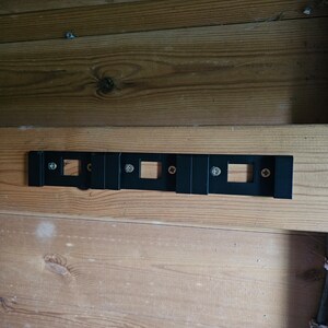May include: A black metal wall mount with five rectangular openings and four screws. The mount is attached to a wooden wall.