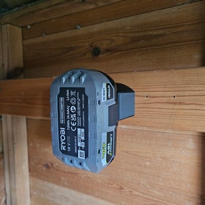 Op de afbeelding: Een grijze en gele Ryobi 18V 4.0Ah lithium-ion batterij met de tekst "Ryobi One+ 18V Lithium+ 4.0Ah 72Wh (4.0Ah) CE Li-Ion" gedrukt op de zijkant. De batterij is gemonteerd op een houten muur.