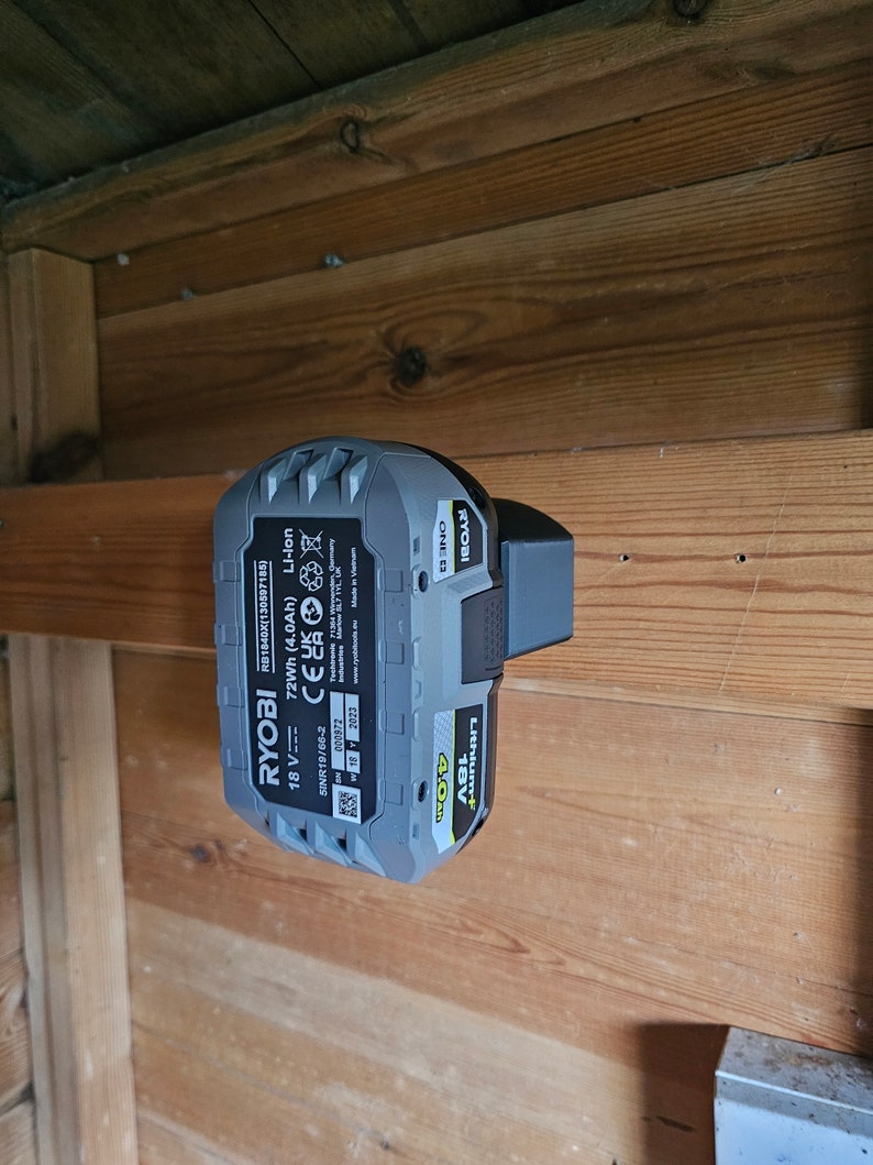 Op de afbeelding: Een grijze en gele Ryobi 18V 4.0Ah lithium-ion batterij met de tekst "Ryobi One+ 18V Lithium+ 4.0Ah 72Wh (4.0Ah) CE Li-Ion" gedrukt op de zijkant. De batterij is gemonteerd op een houten muur.