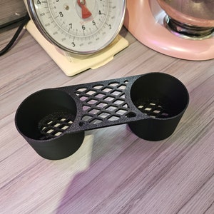 May include: A black, 3D-printed cup holder with two circular openings and a diamond-patterned bridge. The holder sits on a light-colored wooden surface. A vintage kitchen scale and a pink stand mixer are in the background.