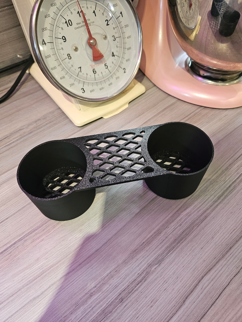 May include: A black, 3D-printed cup holder with two circular openings and a diamond-patterned bridge. The holder sits on a light-colored wooden surface. A vintage kitchen scale and a pink stand mixer are in the background.