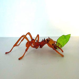 May include: A detailed, amber-colored ant sculpture carrying a bright green leaf. The ant has a spiky back and long, slender legs. The leaf is a vibrant green, contrasting with the ant's body. This unique piece could be a decorative item or a whimsical art piece.