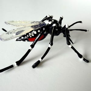 以下が含まれることがあります： 光沢のある黒い体に白い斑点、白い帯が入った長い黒い脚、半透明の羽を持つ蚊のガラス彫刻。腹部には鮮やかな赤色のアクセントがあり、全体的に細部まで作り込まれた写実的な形状をしています。