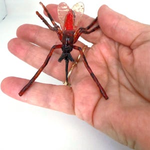 May include: A detailed, handcrafted insect sculpture with a red body, transparent wings, and long, slender legs. The insect is made of a translucent material, with dark brown and black accents. The sculpture is held in a hand.