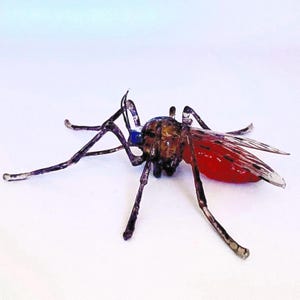Puede incluir: Una escultura de vidrio de un mosquito con abdomen rojo y patas negras. El mosquito tiene una cabeza azul y negra y está posado con las patas extendidas.