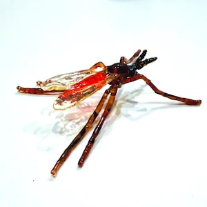 Puede incluir: Una escultura detallada de un insecto con alas transparentes, abdomen rojo y patas marrones. El cuerpo del insecto es de color marrón oscuro y las alas tienen una calidad clara e iridiscente. La escultura está sobre un fondo blanco.