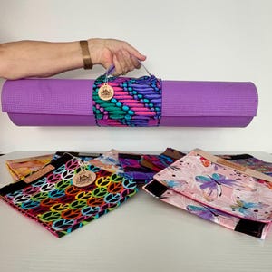 Puede incluir: Una esterilla de yoga morada enrollada, asegurada con una correa estampada colorida con diseños en rosa, azul y morado. Debajo, se exhiben varias bolsas para esterillas de yoga con diversos diseños y colores.