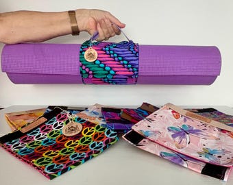 Può includere: Un tappetino da yoga viola fissato con una cinghia in tessuto a motivi ovali blu, rosa e verdi. Sotto ci sono bustine in tessuto piegate, una con un motivo colorato di simboli della pace e l'altra con una stampa di farfalle rosa chiaro.