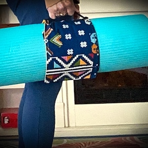 May include: A rolled-up turquoise yoga mat secured with a patterned fabric strap. The strap features a navy blue background with colorful geometric and floral designs. The mat is being carried.