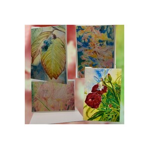 May include: Four watercolor greeting cards featuring nature scenes. One card shows a single leaf with a fairy sitting on it. Another card shows a hummingbird sitting on a red flower. The third card shows a cluster of pink flowers. The fourth card shows a blue and yellow abstract design.