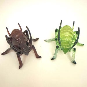 Puede incluir: Dos figuras de insectos de vidrio. Una es de color marrón translúcido y la otra de color verde translúcido. Ambos tienen patas, antenas y segmentos corporales detallados. Los insectos se muestran sobre un fondo blanco.