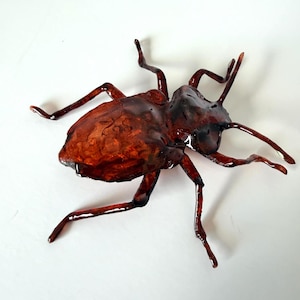 May include: A glossy, dark amber-coloured beetle figurine with a translucent, textured shell and six spindly legs. The insect sculpture features deep reddish-brown tones and a smooth, reflective surface that catches the light, highlighting its segmented body and antennae.