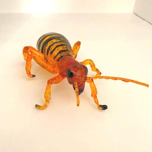 May include: A detailed, orange and black insect sculpture with a segmented body and long antennae. The insect has a reddish-brown head and legs, and the body is striped with black and orange. The sculpture is set against a white background.