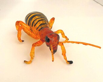 Könnte beinhalten: Detaillierte, orange und schwarze Insektenskulptur mit einem segmentierten Körper und langen Antennen. Das Insekt hat einen rotbraunen Kopf und Beine, und der Körper ist schwarz und orange gestreift. Die Skulptur steht vor weißem Hintergrund.