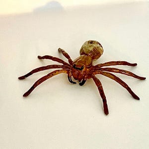 May include: A detailed, brown spider figurine with eight legs and a rounded body. The spider has black accents on its fangs and eyes, and the body has a textured appearance. The figurine is set against a plain white background.