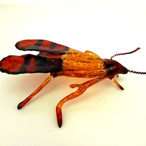 May include: A decorative insect figurine crafted with a translucent amber-colored body and head. The wings are a deep reddish-brown with distinct black horizontal stripes. It features two thin, textured antennae and slender, jointed legs.