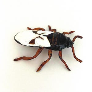 Puede incluir: Figura artesanal de insecto con diseño de alas blancas y negras, cuerpo negro y patas marrones. El insecto está representado en un estilo realista, con detalles y un acabado brillante. La figura está sobre un fondo blanco.