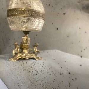 May include: A vintage, ornate, gold-toned metal mirror with a distressed, aged finish. The mirror has a decorative, three-legged stand with a round top. The mirror has a textured surface with small, dark spots.