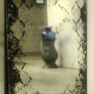 May include: A vintage-style mirror with a distressed black frame and a decorative etched pattern around the edges. The mirror reflects a blurry image of a vase and a room.
