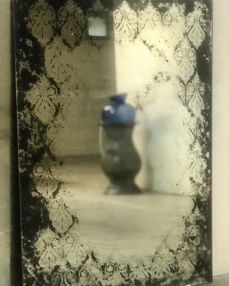 May include: A vintage-style mirror with a distressed black frame and a decorative etched pattern around the edges. The mirror reflects a blurry image of a vase and a room.