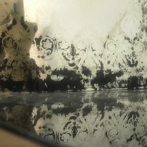 May include: A close-up of a vintage mirror with a distressed, aged look. The mirror has a decorative, ornate border with a floral pattern. The mirror is reflecting the surrounding environment, creating a hazy, blurred effect.