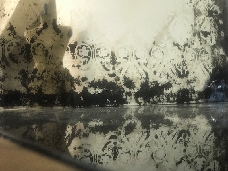 May include: A close-up of a vintage mirror with a distressed, aged look. The mirror has a decorative, ornate border with a floral pattern. The mirror is reflecting the surrounding environment, creating a hazy, blurred effect.