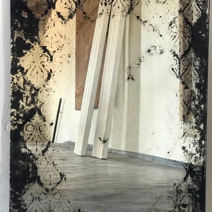 May include: A large, rectangular mirror with a distressed, black-painted frame. The frame features a decorative, gold-colored pattern. The mirror is reflecting a room with white walls and wooden beams.