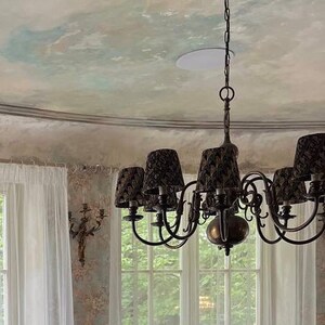 May include: A black chandelier with multiple lights hangs from a decorative ceiling medallion. The chandelier has a dark metal frame and black fabric shades. The ceiling is painted in a light gray color with a subtle blue and green marbling effect.