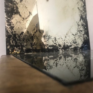 May include: A vintage mirror with a distressed, aged look. The mirror has a decorative border with a floral pattern. The mirror is reflecting the surface it is sitting on.