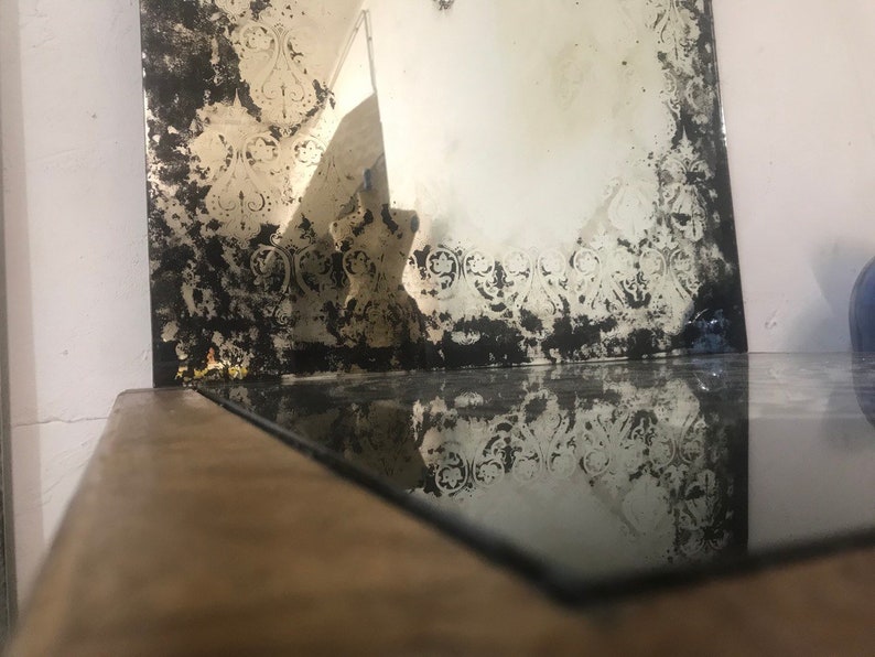May include: A vintage mirror with a distressed, aged look. The mirror has a decorative border with a floral pattern. The mirror is reflecting the surface it is sitting on.