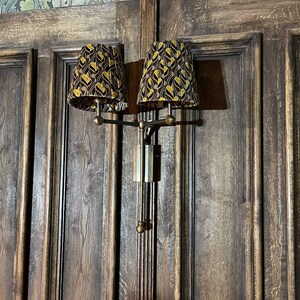 May include: A pair of wall-mounted lamps with brown wooden shades featuring a geometric pattern in gold and black. The lamps are mounted on a brass arm and have a dark wood background.