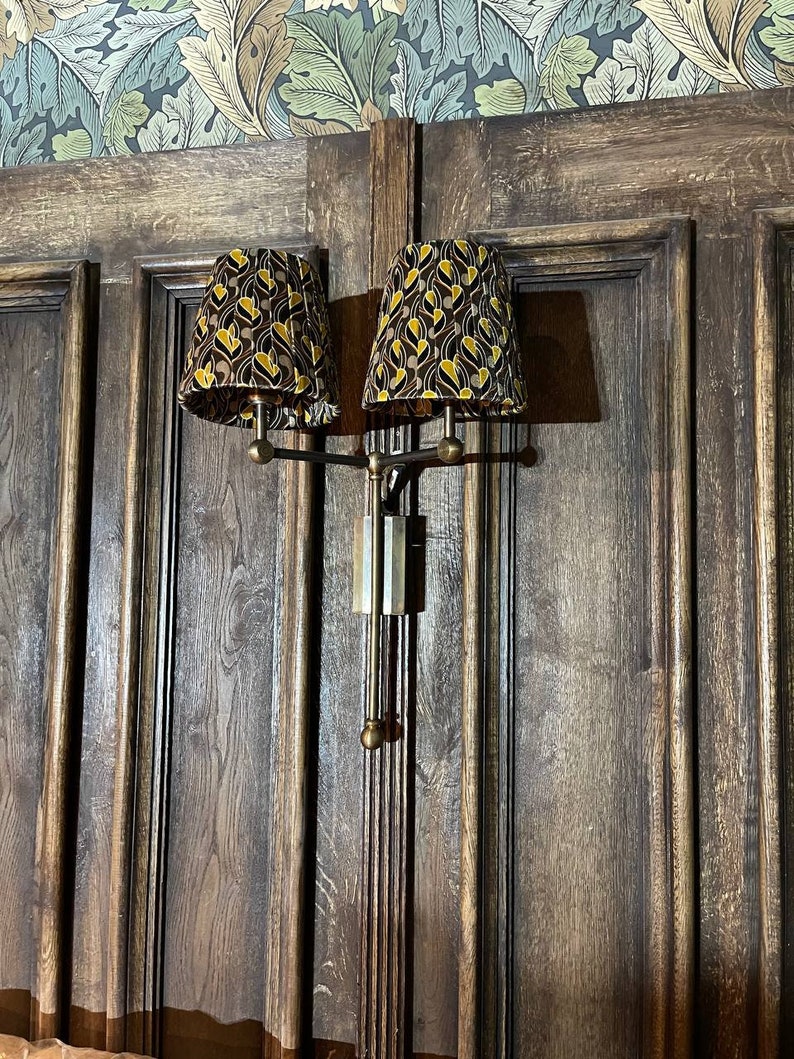 May include: A pair of wall-mounted lamps with brown wooden shades featuring a geometric pattern in gold and black. The lamps are mounted on a brass arm and have a dark wood background.