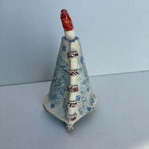 May include: A ceramic sculpture with a cone-shaped body, decorated with a blue and white floral pattern. It has a red, textured top and red and white accents. The base has three small feet. The sculpture is likely handmade.